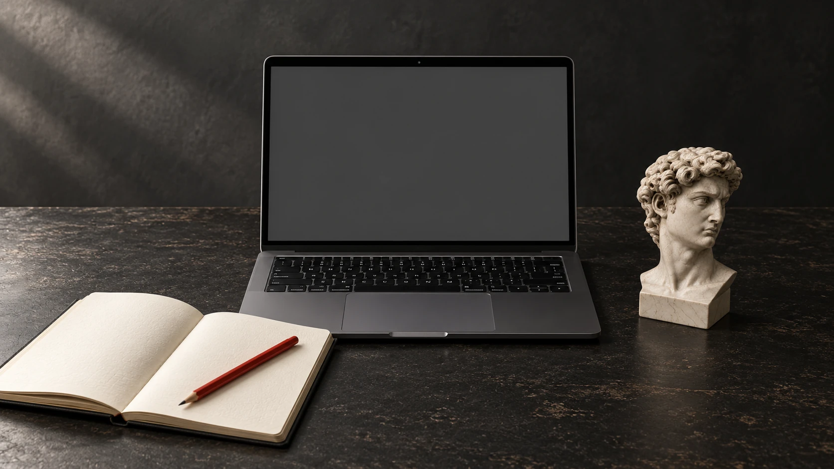 Escritorio con laptop, cuaderno, lápiz rojo y un busto del David